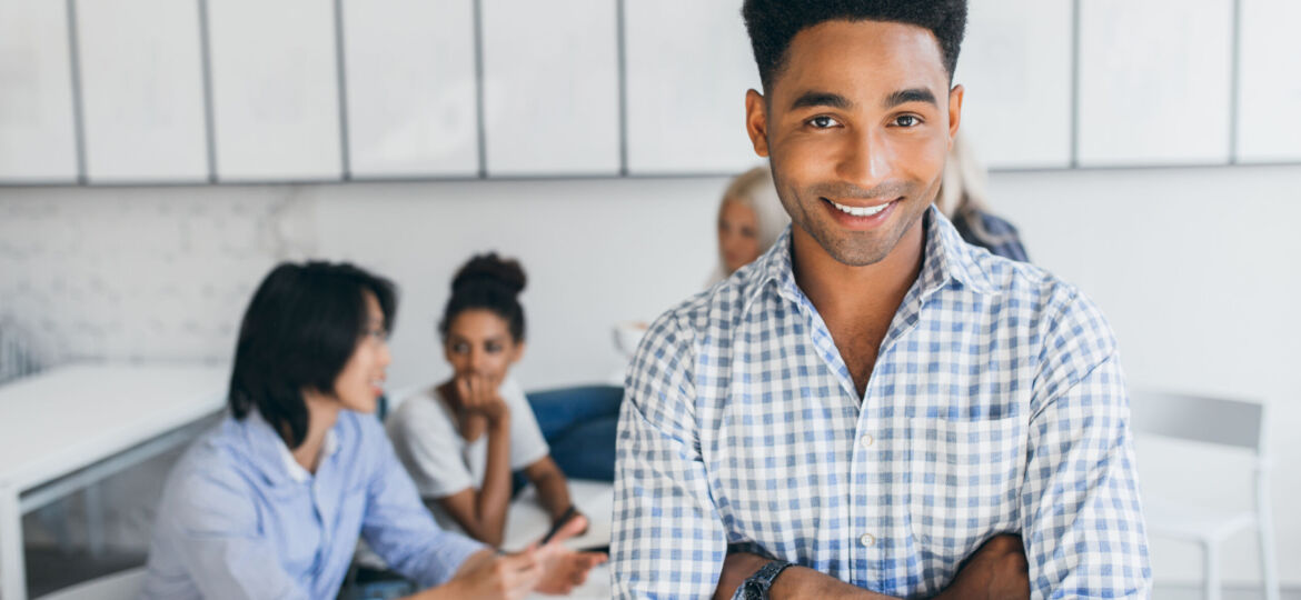 Glad young man with african hairstyle posing with arms crossed in his office with other employees on background. Male manager in blue shirt smiling during conference at workplace..