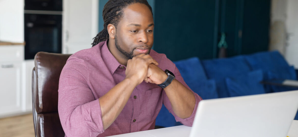 Focused and concerned black man in smart casual shirt looks at laptop screen