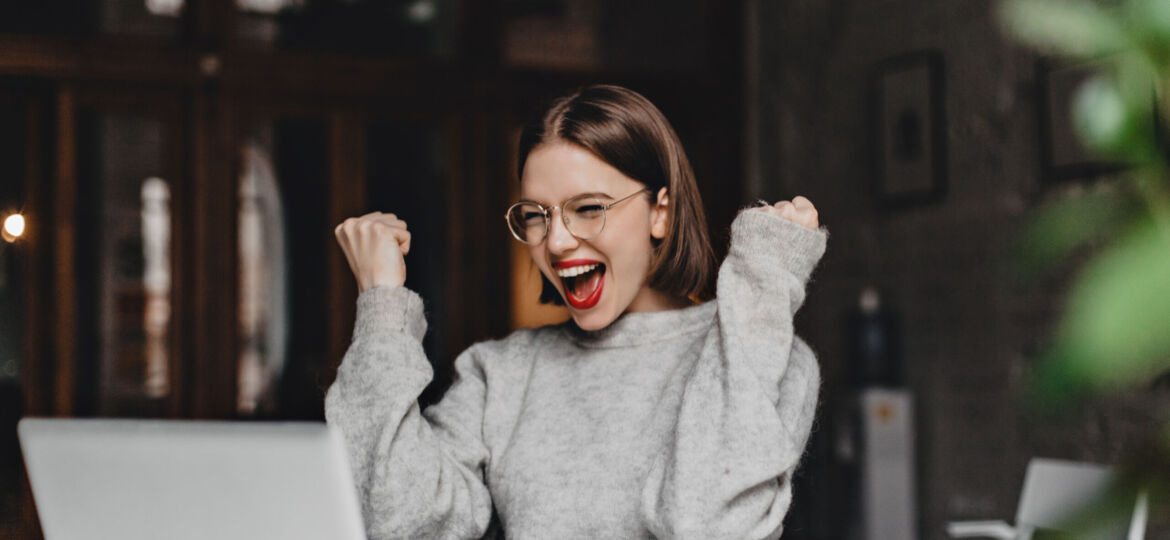 Happy woman in glasses makes winning gesture and sincerely rejoices. Lady with red lipstick dressed in gray sweater looking at laptop.