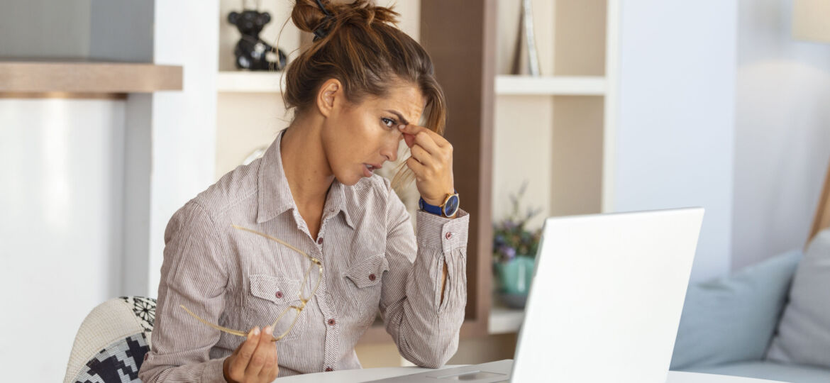 Young frustrated woman working at office desk in front of laptop suffering from chronic daily headaches,