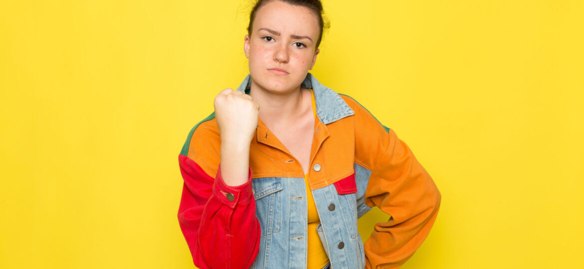 front-view-young-female-yellow-shirt-colorful-jacket-blue-jeans-posing-showing-her-fist