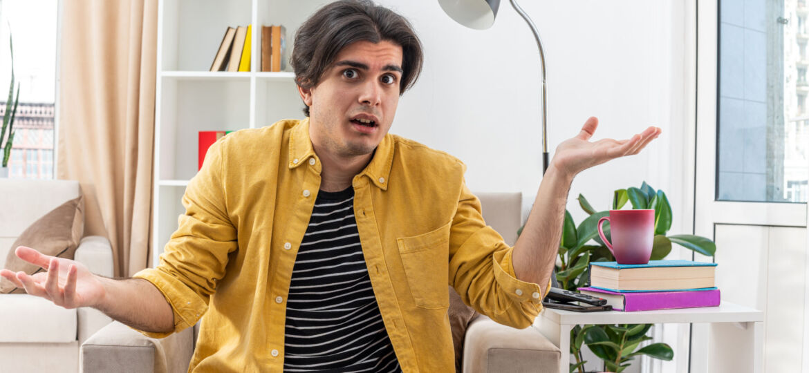 young man in casual clothes looking at camera confused spreading arms to the sides sitting on the chair in light living room