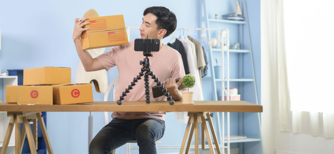 An Asian man is showing clothes in front of smartphone  live streaming at his shop. Technology online business concept.