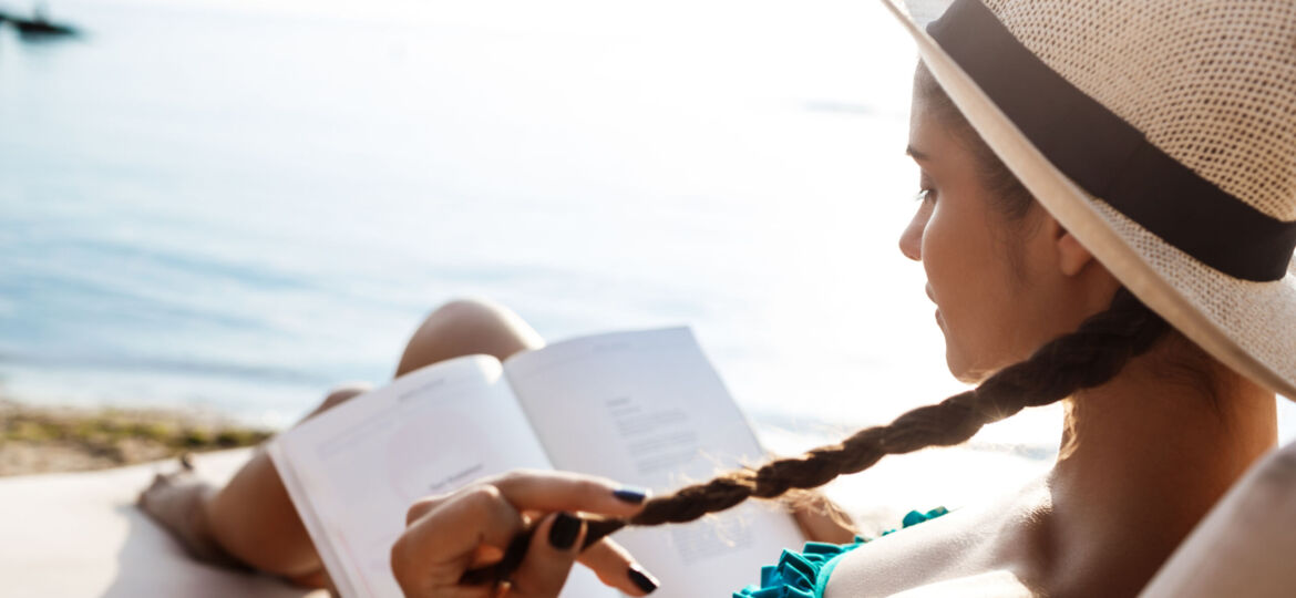 Beautiful brunette girl in hat reading book, lying at beach.