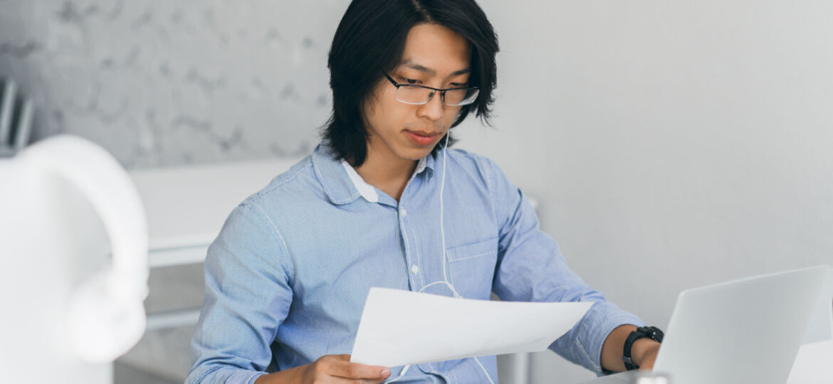 Concentrated asian office worker in earphones reading documents at workplace. Indoor portrait of chinese freelance it-specialist drinks coffee while using with laptop..