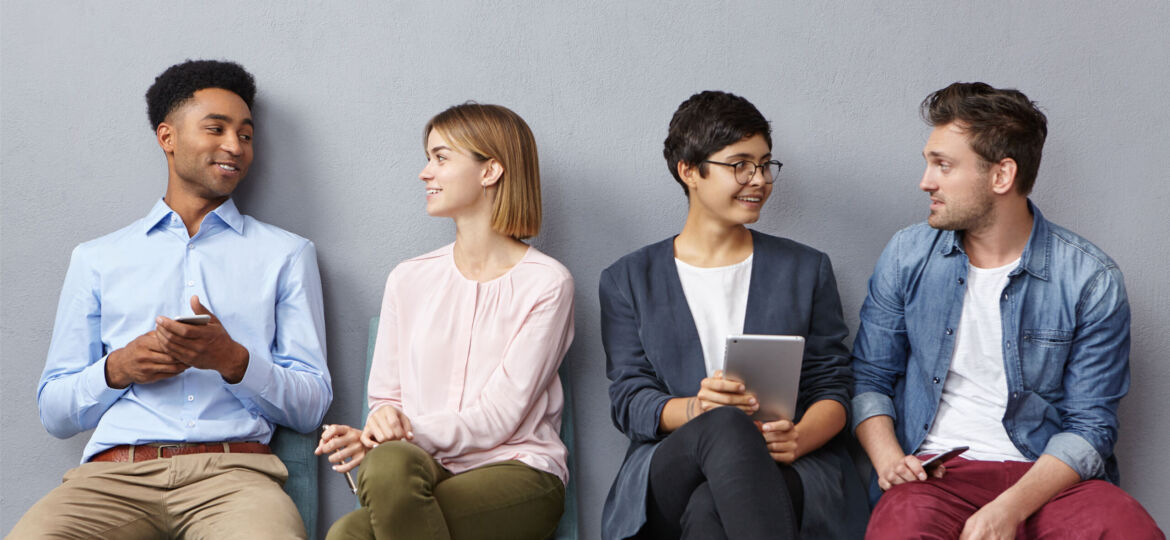 Horizontal portrait of people sit in queue, have pleasant conversation with each other, share ideas and life experience, isolated over grey concrete wall. Diverse group in row, speak and hold gadgets