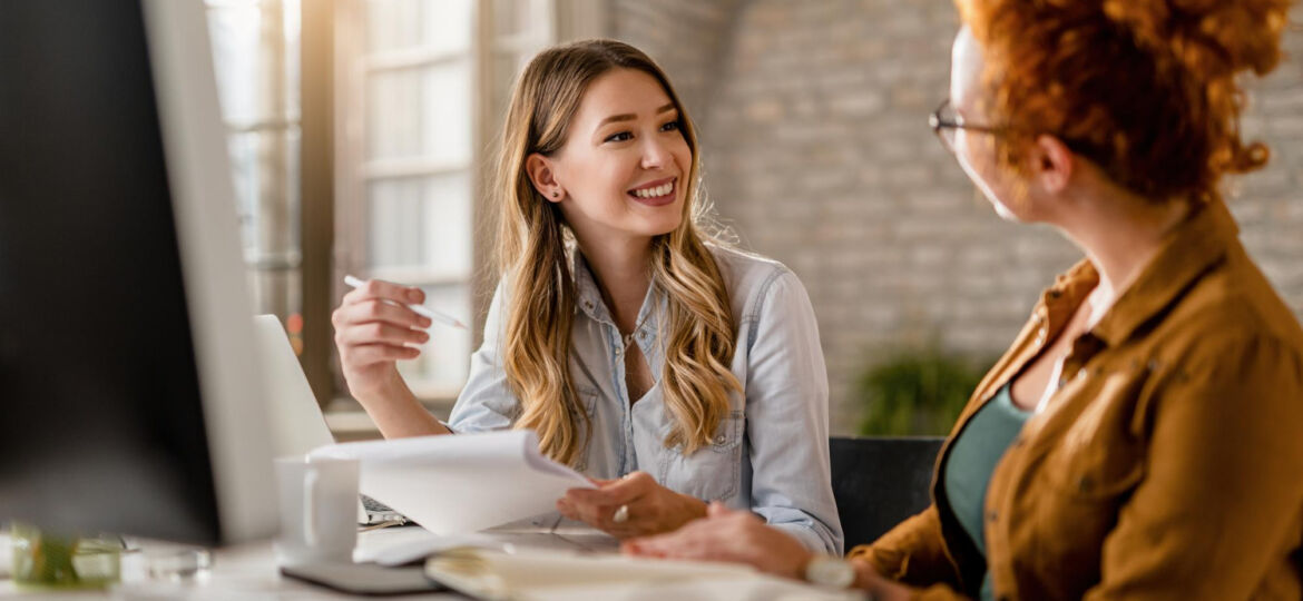 smiling-creative-businesswoman-going-through-paperwork-talking-her-female-colleague-office