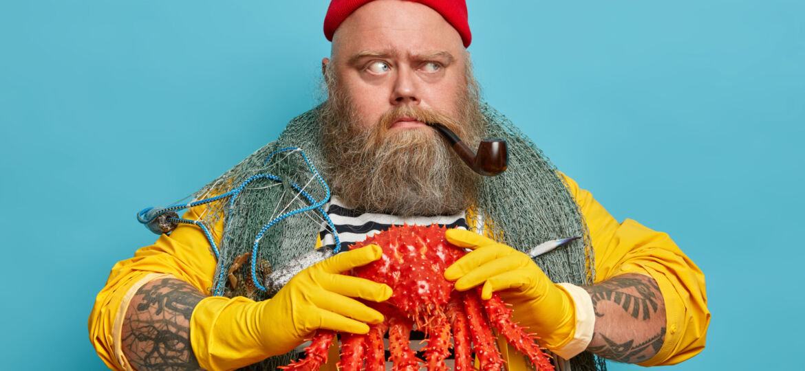 Photo of serious bearded plump seaman assists during voyage, goes fishing, smokes pipe and looks with angry expression aside, works in navy, holds big octopus, stands against blue background.
