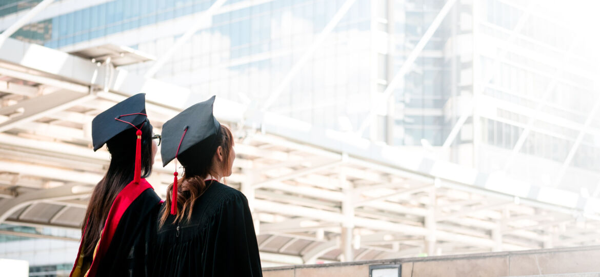 Two girls in black gowns standing look up to the sky with happy graduates.