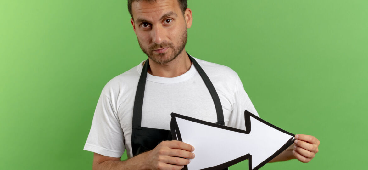 barber man in apron holding white arrow looking at camera with serious face standing over green background