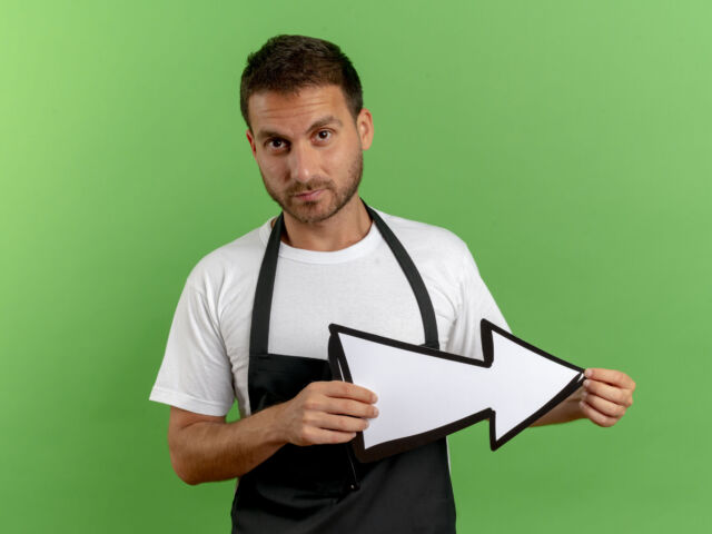 barber man in apron holding white arrow looking at camera with serious face standing over green background