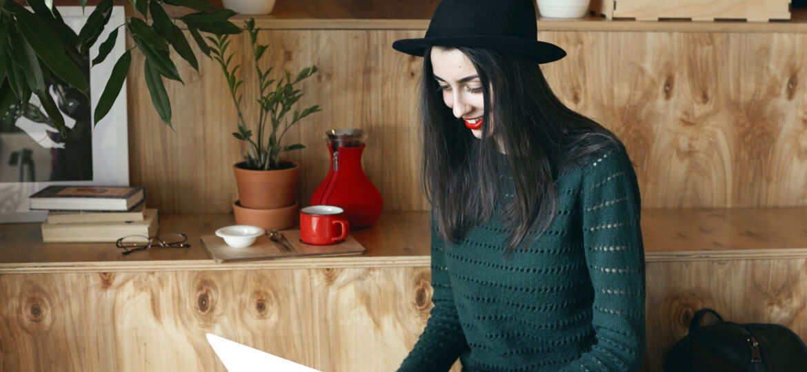Charming young woman with long dark hair is smiling at the screen of a laptop in a cafe. Attractive female freelancer check e-mail on her notebook against the background of a wooden wall with plants