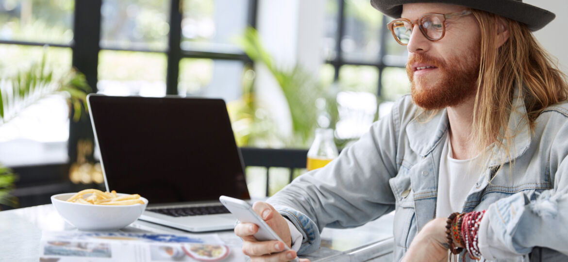 Bearded hipster guy in eyewear and hat, reads text message on smart phone, sits in spacious room, reads magazines, uses laptop computer. Fashionable copywriter checks work of new application
