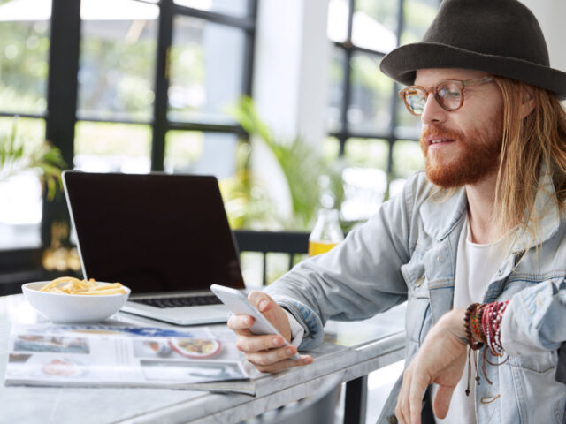 Bearded hipster guy in eyewear and hat, reads text message on smart phone, sits in spacious room, reads magazines, uses laptop computer. Fashionable copywriter checks work of new application