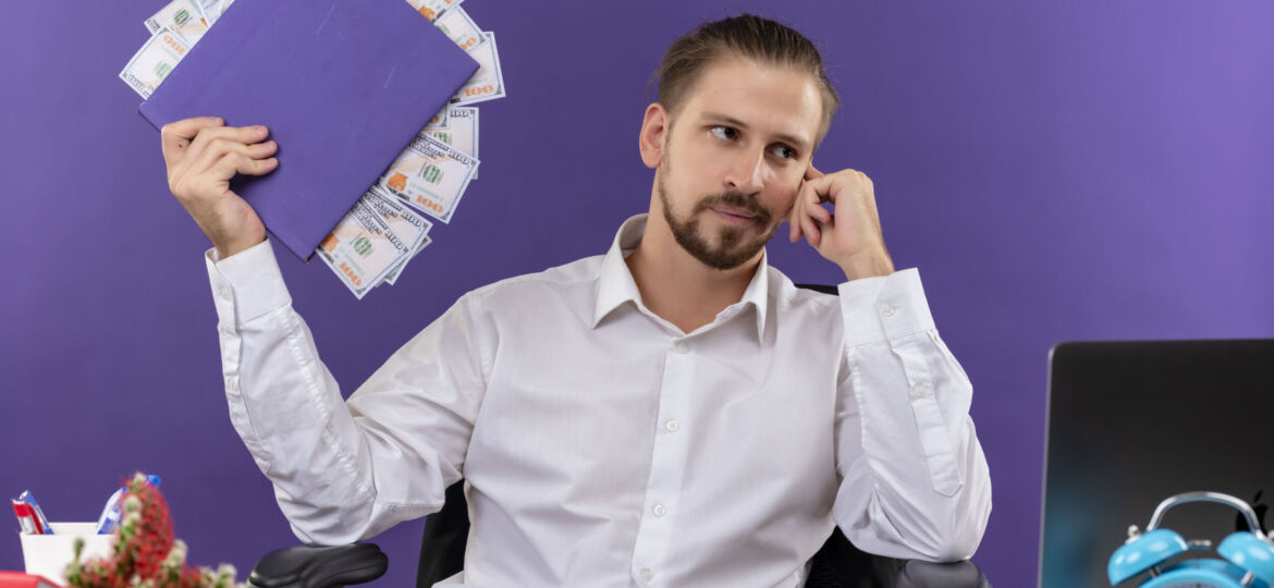handsome businessman in white shirt holding folder with cash looking aside with pensive expression sitting at the table in offise over purple background