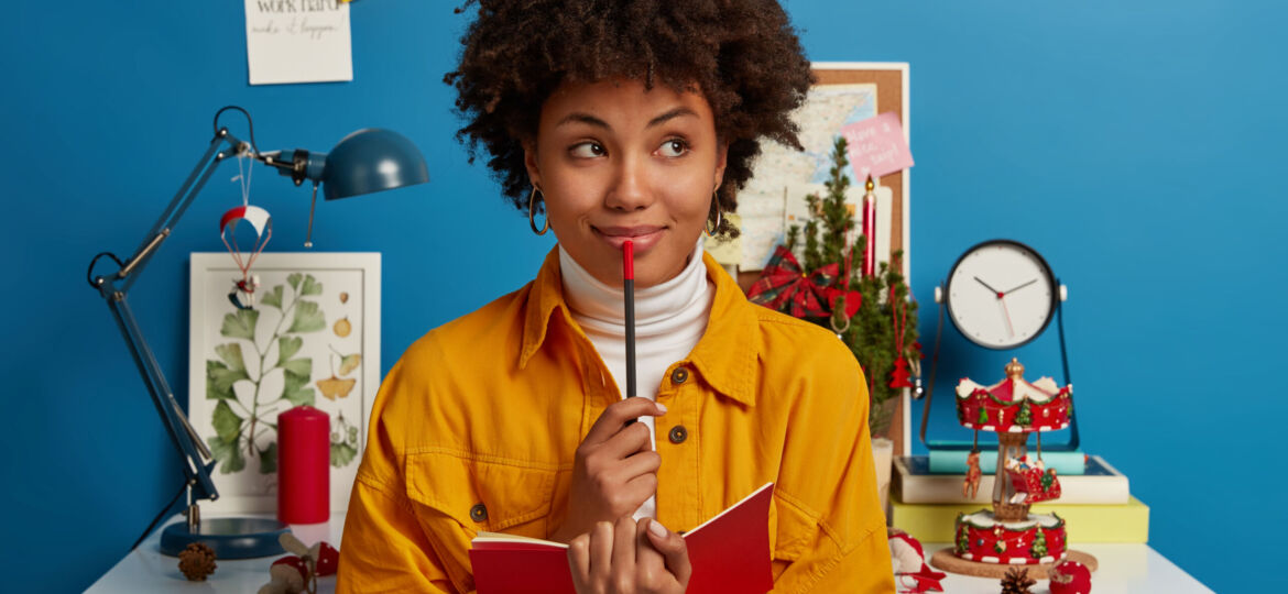 Pensive dark skinned girl creats interesting composition, sits near workplace with opened notebook and pencil, looks pensively aside, does hometask, prepares for examination, isolated on blue wall