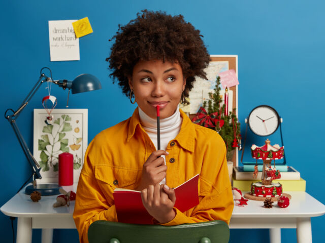 Pensive dark skinned girl creats interesting composition, sits near workplace with opened notebook and pencil, looks pensively aside, does hometask, prepares for examination, isolated on blue wall
