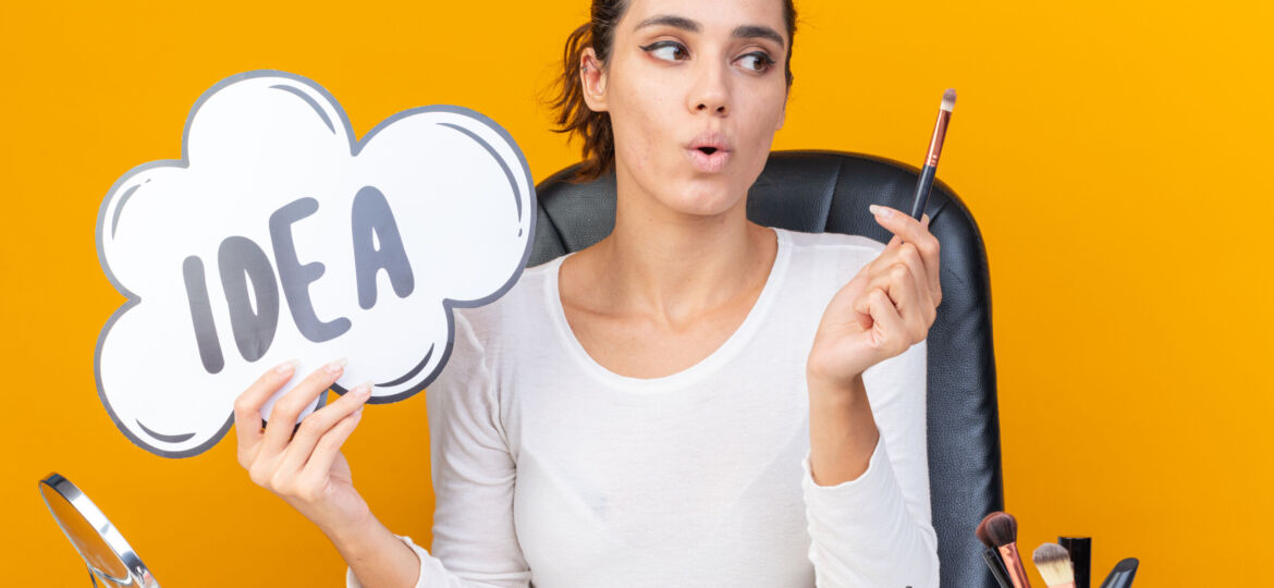 surprised-pretty-caucasian-woman-sitting-table-with-makeup-tools-holding-idea-bubble-looking-makeup-brush