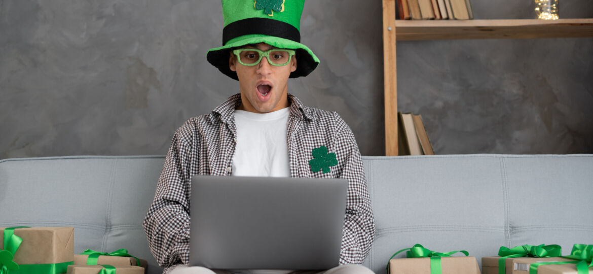 Young arabic man holding laptop scared and amazed with open mouth for surprise, disbelief face at home. St Patricks day online shopping