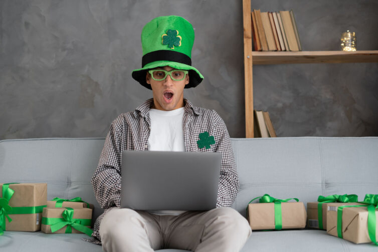 Young arabic man holding laptop scared and amazed with open mouth for surprise, disbelief face at home. St Patricks day online shopping