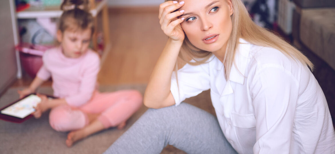 Young mother is worried and sad at home on the floor, the child is watching cartoons on the tablet and playing, a naughty child, family concept.