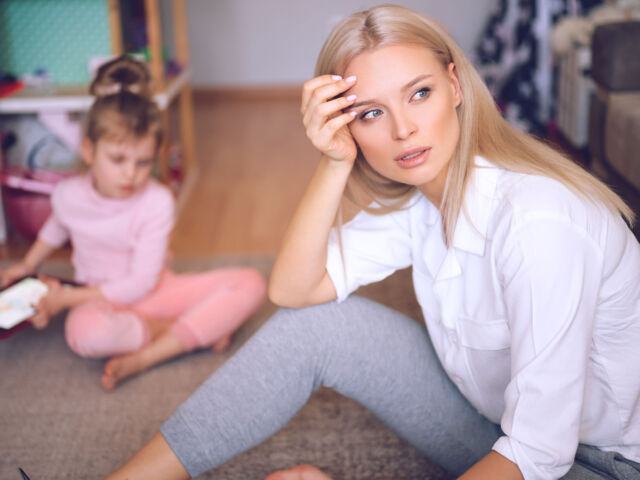 Young mother is worried and sad at home on the floor, the child is watching cartoons on the tablet and playing, a naughty child, family concept.