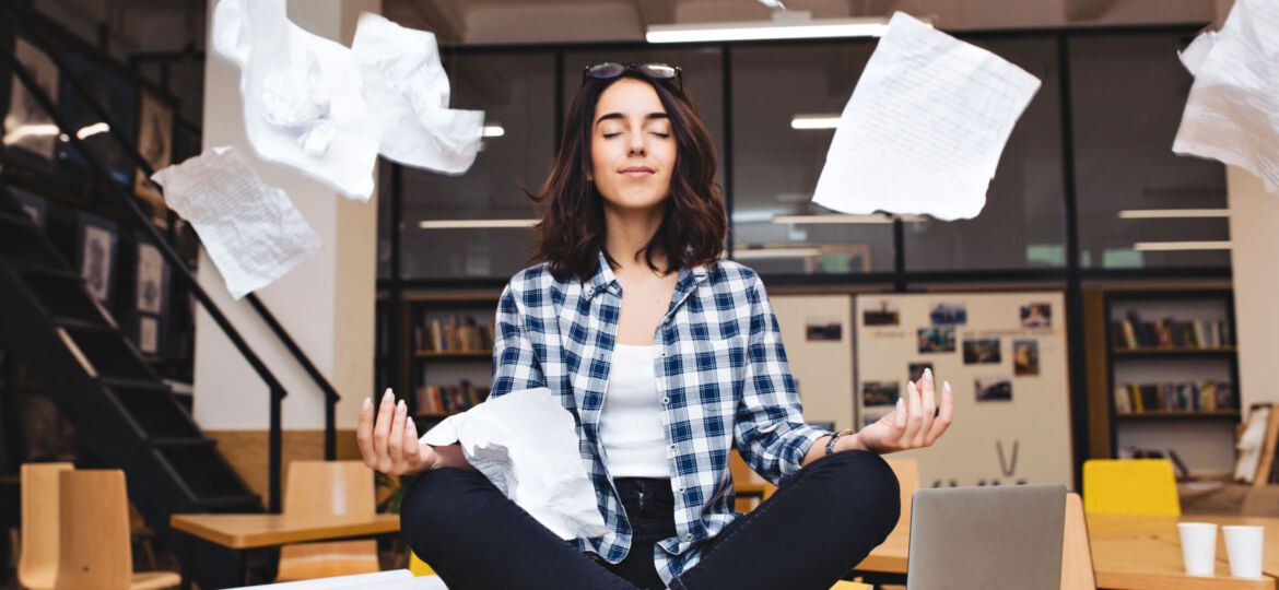 Young pretty joyful brunette woman meditating on table surround work stuff and flying papers. Cheerful mood, taking a break, working, studying, relaxation, true emotions.