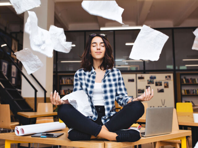 Young pretty joyful brunette woman meditating on table surround work stuff and flying papers. Cheerful mood, taking a break, working, studying, relaxation, true emotions.