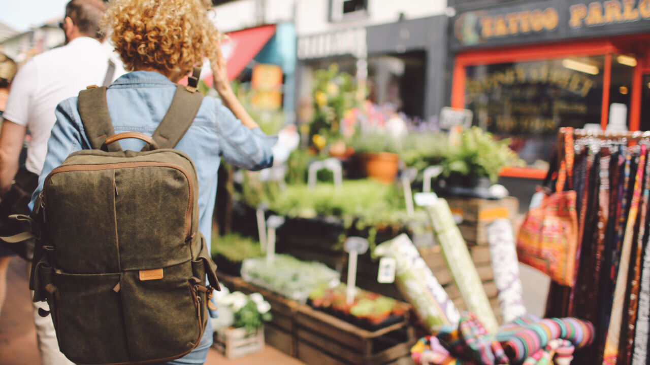 rear-view-woman-walking-through-street-market-stalls-city A person with curly hair wearing a denim jacket and olive-green backpack walks through a lively outdoor market lined with colourful fabric rolls, plants, and local shops, capturing the spirit of shopping local in a vibrant community.
